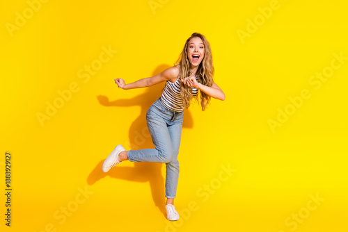 Excited young woman in trendy casual outfit leaping and smiling against vibrant yellow background