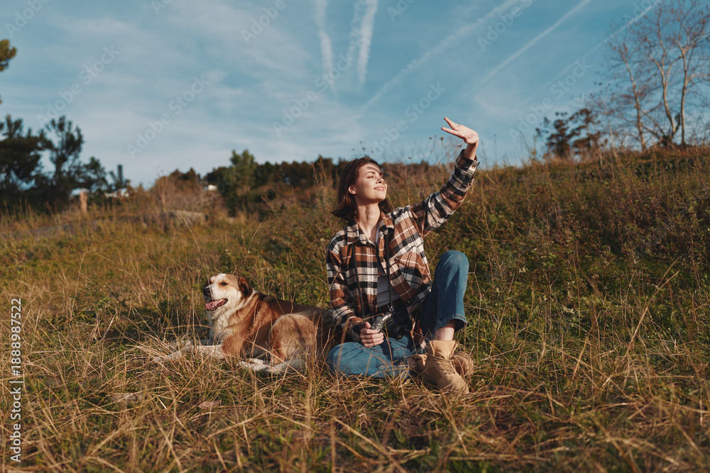 Fototapeta premium Woman and dog in an open field under a blue sky, enjoying nature and sunshine. Casual plaid shirt, jeans and boots as she sits and waves toward the bright horizon.