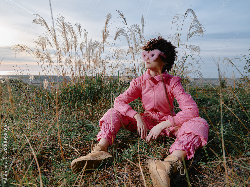 Fototapeta premium Outdoor fashion scene with a model wearing a pink jumpsuit and mask, seated in tall grasses during golden hour, presenting bold style and nature inspired calm.