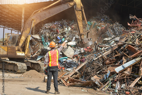 Rear view of a worker at a metal scrap yard pointing at an excavator with a magnet, Thailand