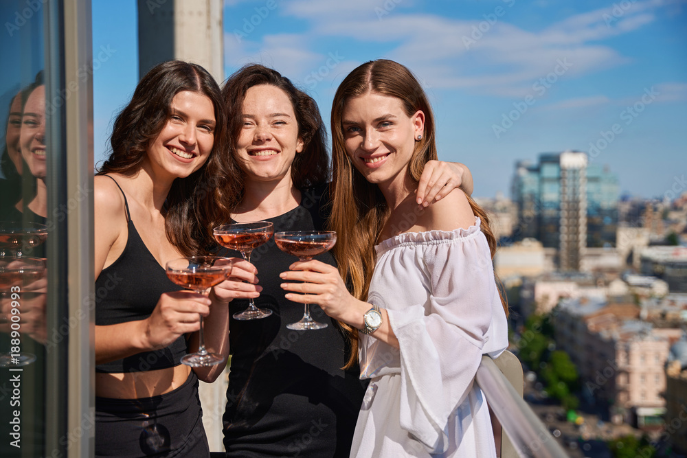 Fototapeta premium Smiling young women posing with glasses of champagne on terrace outdoors