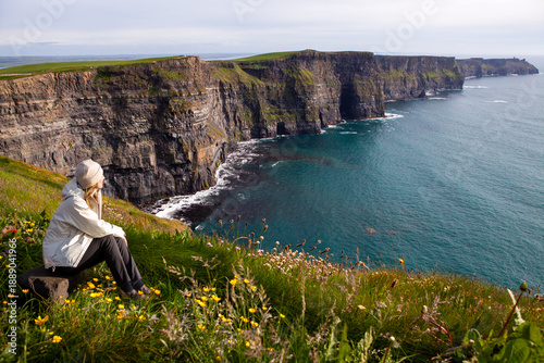 A solitary traveler sits amidst yellow wildflowers on a lush green cliffside, gazing out at the dramatic, rugged coastline of Cliffs of Moher and the vast blue Atlantic Ocean.