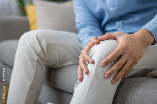 Tableau sur toile Middle-aged man sitting on a couch pressing his painful knee, grimacing from joi