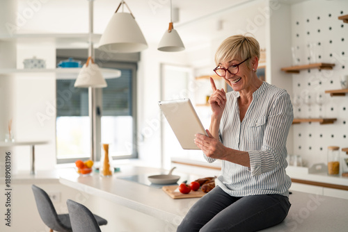 Fotografi Senior Woman Smiling and Working on Tablet in Bright Modern Kitchen at Home