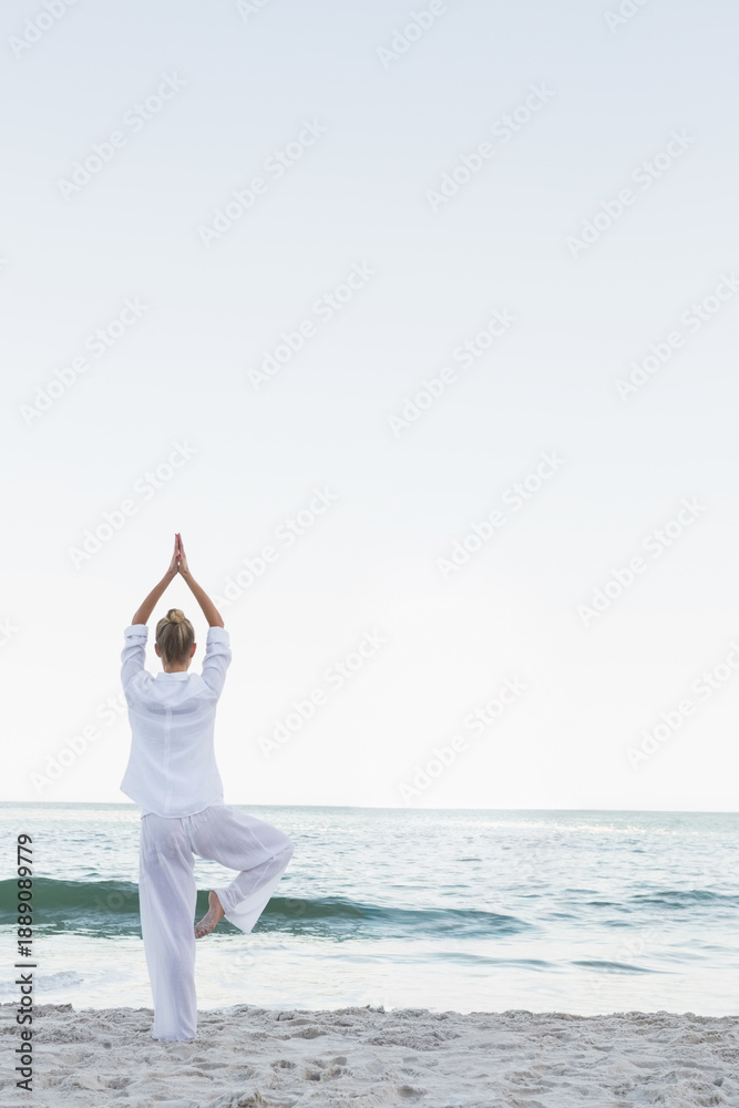 Fototapeta premium Woman is holding yoga tree pose on sandy shoreline, wearing loose white clothing, copy space