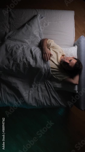 Vertical video man lies awake in bed at night under dim light, restless and anxious, struggling with insomnia and sleeplessness while staring at the ceiling, alone and exhausted