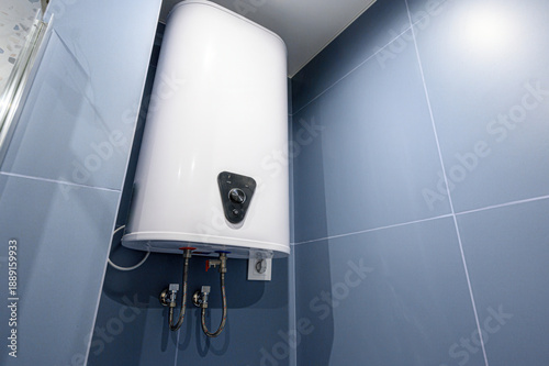 White electric water heater mounted on blue tiled wall in bathroom, with control knob, water inlet outlet pipes, and electrical connection visible.