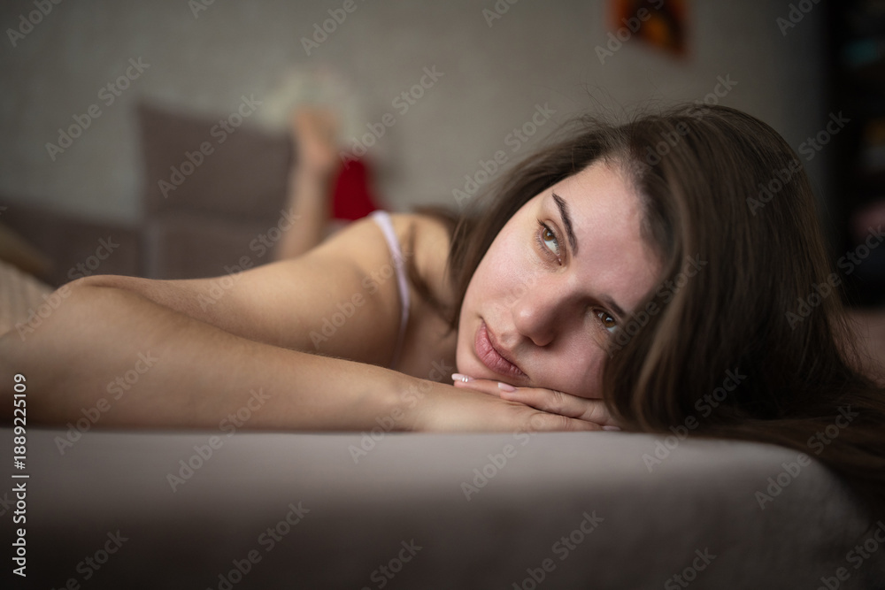 Obraz premium Young woman with long dark hair resting on a couch, wearing a white shirt, with soft pillows and a neutral background creating a cozy indoor atmosphere
