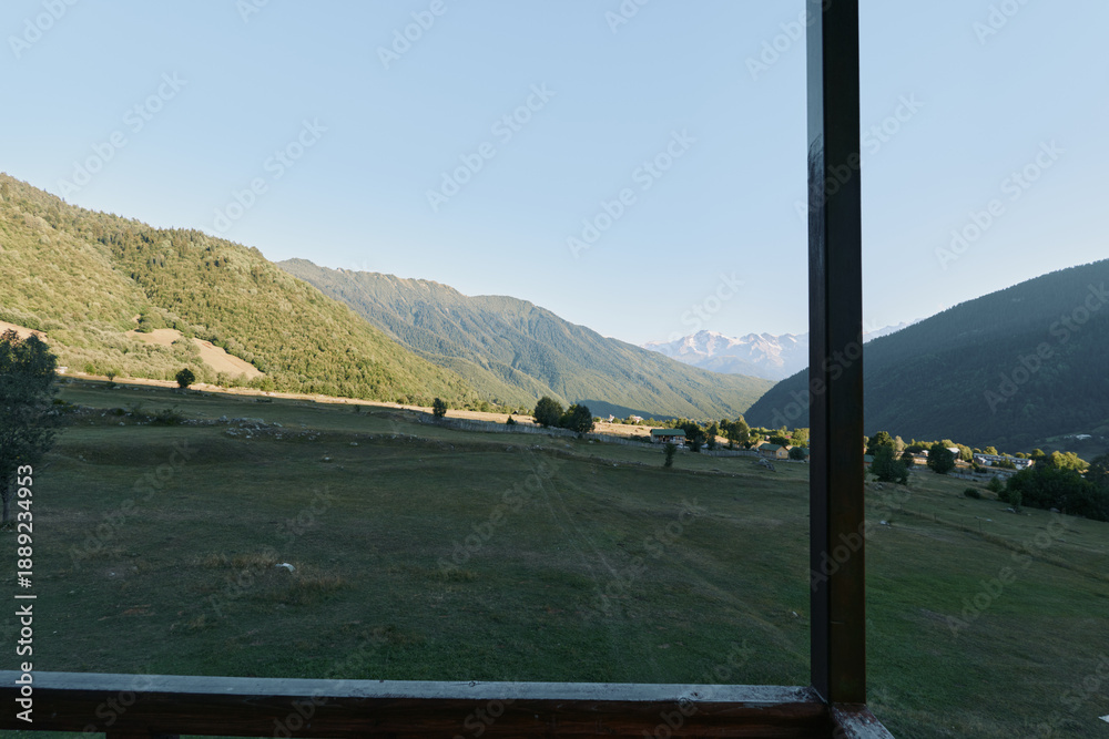 Obraz premium A serene landscape is seen through a window, revealing rolling valley plains and distant mountains under a clear sky. The window frame adds depth, inviting quiet contemplation of nature’s calm beauty.