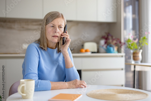 Принти на полотні Elderly woman in kitchen listens intently on her mobile phone, worried and pensi