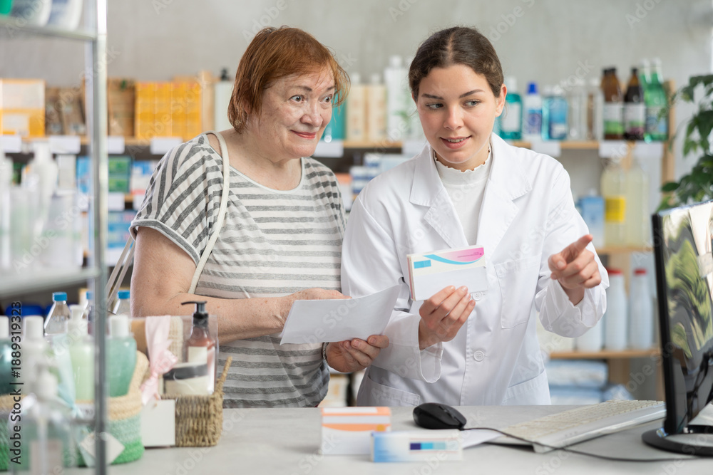 Obraz premium Young woman pharmacist looking for prescription on computer to elderly woman buyer in pharmacy