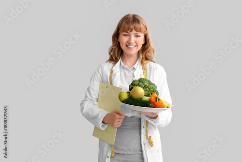 Beautiful young happy female nutritionist with vegetables, clipboard and meas...