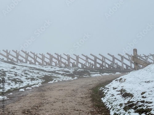 Snowy path leads to a wooden fence on a foggy day in the countryside with limited visibility