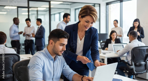 Businesswoman guiding colleague on laptop in modern office setting with team working in background