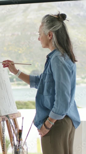 Vertical video: Sunlight bathing senior painter at easel using thin brush, capturing view, denim