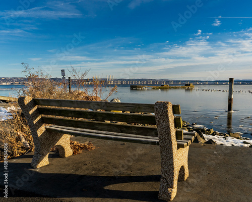 Nyack, NY - US - Jan 24, 2026 Serene winter waterfront in Nyack, NY, with a concrete bench overlooking a partly frozen Hudson River and the Mario M. Cuomo Bridge beneath a bright blue sky.