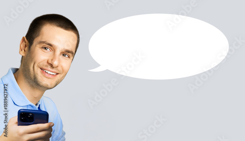 Portrait image - happy smile young man wear blue t-shirt cloth, holding hand cell phone mobile smartphone cellphone, isolated grey gray background. White empty mockup speech bubble with free space