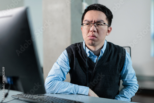 Fotografie Young Asian man looks at computer with a skeptical expression in the office, ideal for AI anxiety, job insecurity, or technology distrust concepts
