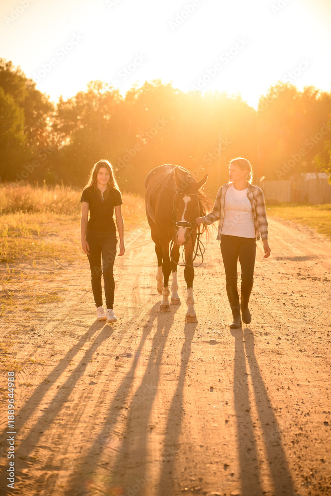 Fototapeta premium Family time in countryside, sunset ride, mother and daughter leading horse by reins along sun-drenched road. Horseback riding in countryside. Caring for and loving horses, equestrian sport.