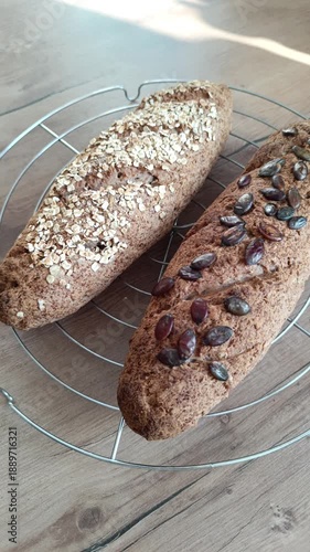 Vertical close-up of two fresh sourdough bread loaves with oats and pumpkin seeds on a cooling rack. Sunlight highlights the crust texture. Ideal for healthy eating, baking, and rustic food concepts.