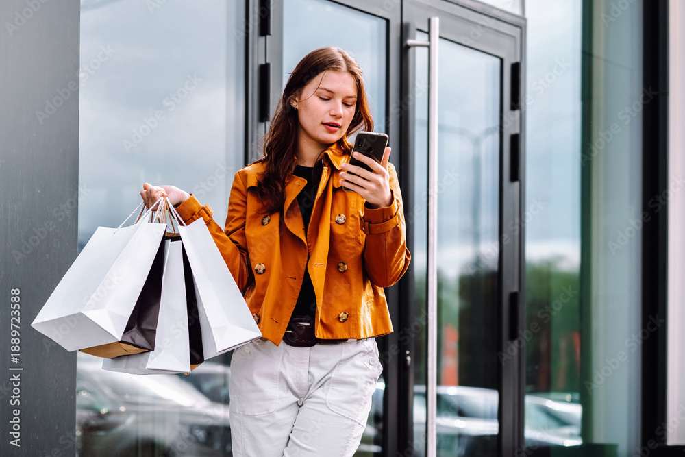 Fototapeta premium Young woman shopping outdoors while checking phone, dressed in casual style with multiple shopping bags. Consumerism, sale, black friday. Online shopping.