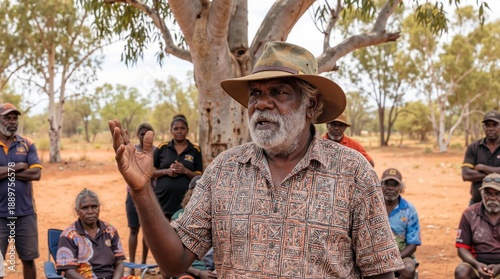 An Aboriginal elder addresses a group of people in a dry, dusty landscape under a tree, sharing knowledge and leading a community discussion. Generative AI.