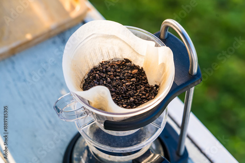 Close-up of used coffee grounds in a paper filter inside a glass drip coffee maker placed outdoors on a sunny day
