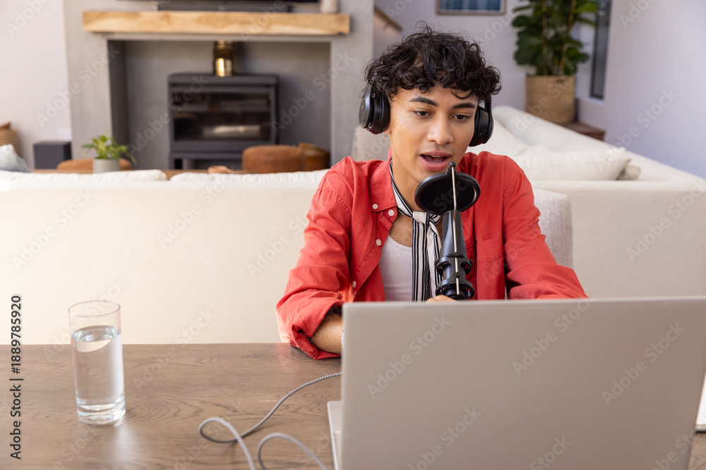 Fototapeta premium Male with headphones speaking into microphone at living room, table with laptop, copy space