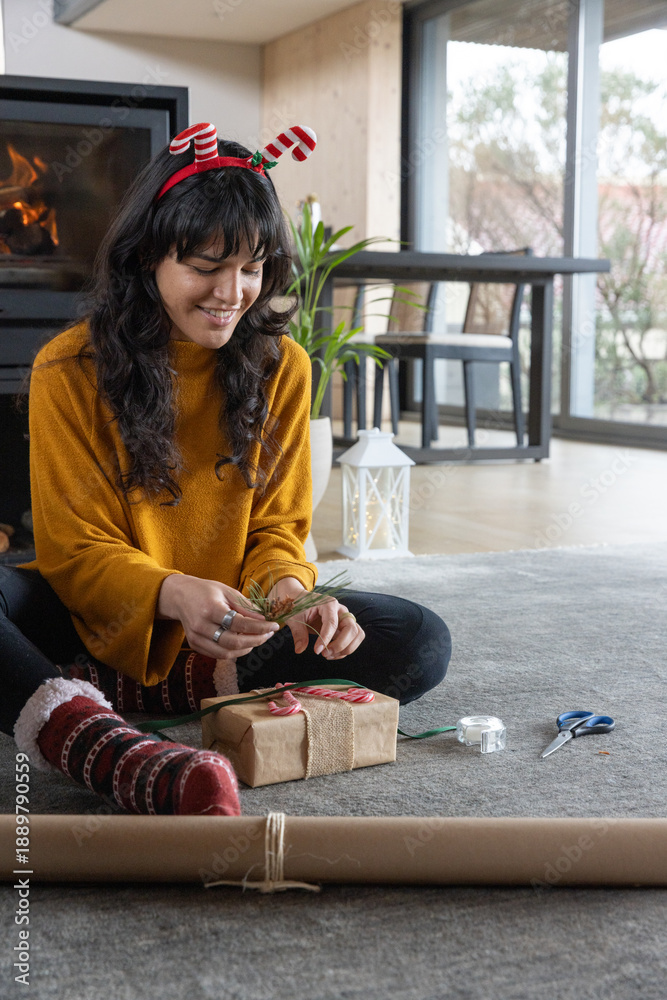 Obraz premium Woman wearing festive headband wrapping presents on rug in living room near fireplace using ribbon