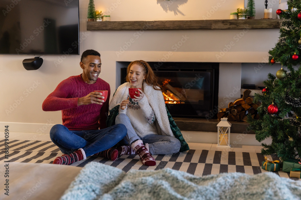 Fototapeta premium Diverse couple sitting on striped rug in front of fireplace with red mugs beside christmas tree