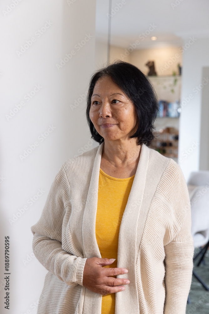 Naklejka premium Senior Asian woman standing inside living room next to column, wearing beige cardigan, mustard top