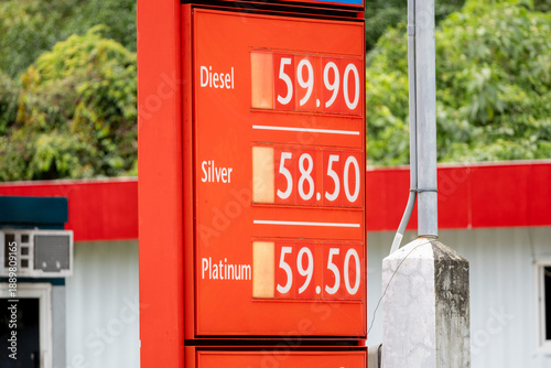 Gas station price board: vivid red display showing fuel prices — diesel at 59.90, Silver grade at 58.50, and Platinum at 59.50, set against a backdrop of lush greenery and a modern gas station structu