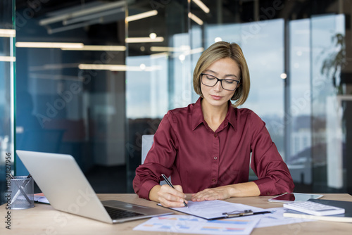 Obraz na plátně Young businesswoman reviewing financial documents and writing notes at desk with