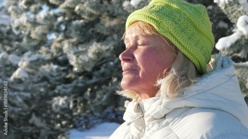 Senior woman with eyes closed breathing in fresh frosty air in snowy winter park on sunny day. Female enjoying serene moment and warmth of sun on face. Sunbathing to produce vitamin D.