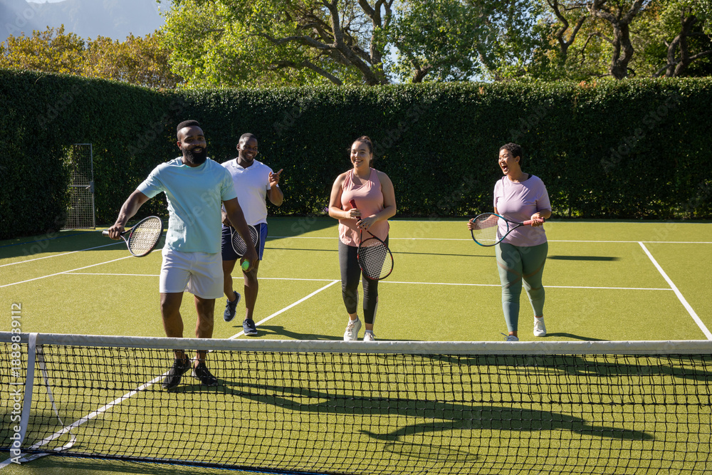 Obraz premium Diverse friends in sportswear walking together on grass court carrying tennis rackets next to net