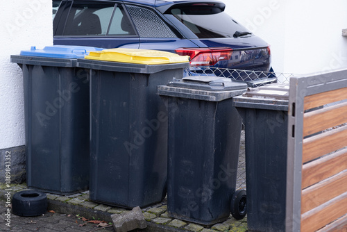 Modern household waste sorting system with black, blue, and yellow lid garbage bins outdoors in a German residential setting near a car.
