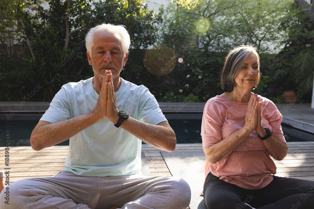 Fototapeta premium Senior couple meditating cross-legged on yoga mats on wooden deck beside pool wearing sport watches