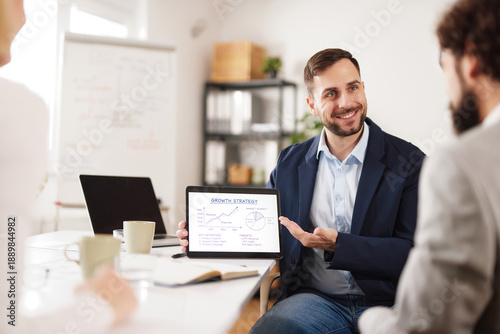 Members of a business team are engaged in a meeting in their office. One person presents a growth strategy on a tablet while others listen and take notes.