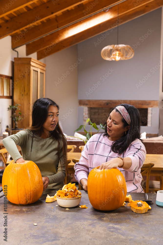 Obraz premium Diverse mother and teen daughter scooping pumpkin pulp and seeds, carving pumpkins in dining room