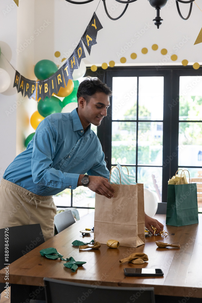 Obraz premium Asian male arranging gift bags on dining table at home beneath happy birthday banner, balloons