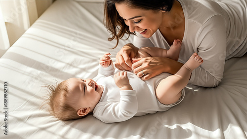Happy mother playing with laughing baby lying on white bed in sunny bedroom enjoying family time and motherhood love together