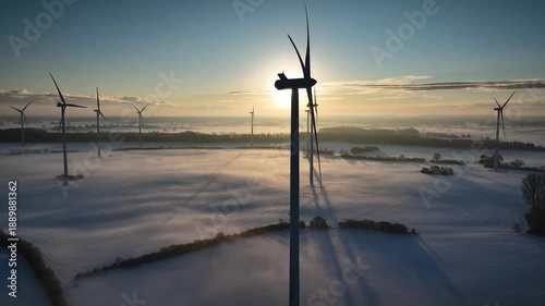 Aerial view of wind farm at sunrise with tall wind turbines across snow-covered agricultural fields in soft morning light and low mist  in calm winter atmosphere. Renewable energy infrastructure.