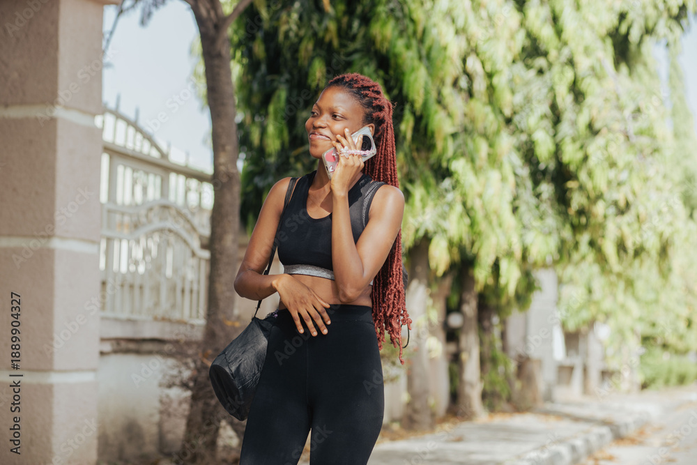 Fototapeta premium Cheerful young girl in sportswear talking by phone smiles at break of yoga at park on sunny day. Black schoolgirl talks with friend by cell phone. Active people..