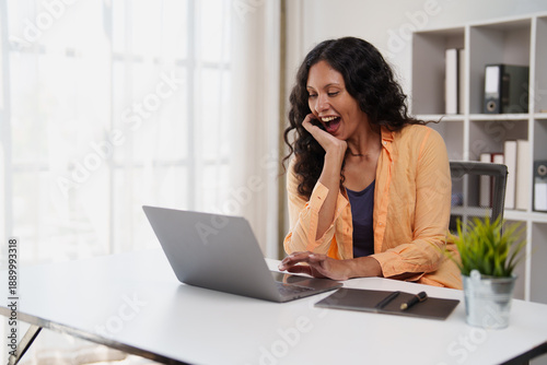 Tableau sur toile Woman with dark curly hair sitting at a white desk, enthusiastically laughing an