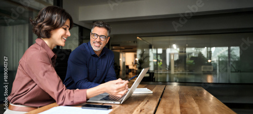 Business team two happy busy executives working together using laptop in office. Young business woman manager showing presentation to mature man investor, consulting client partner at work meeting.