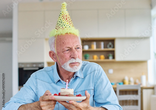 Sad senior elderly man celebreting birthday alone and lonely blowing cake candle sitting at home