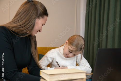 Happy mother supervising her young daughter finishing homework and learning online from home, using a laptop and writing in a notebook, symbolizing family education and homeschooling