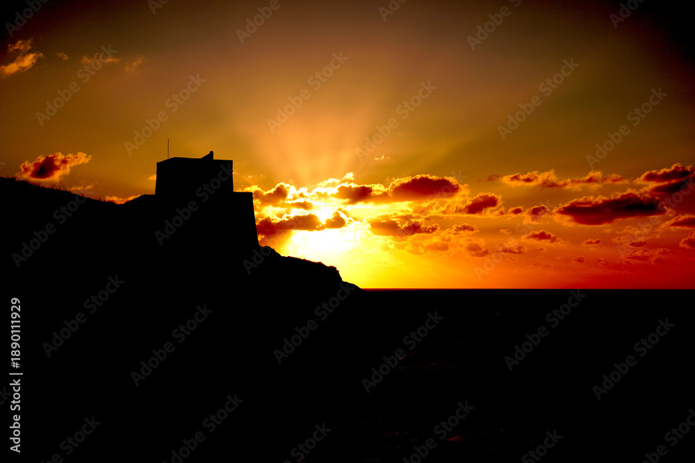 Obraz premium Dramatic silhouette of ancient fortress ruins on hilltop against vibrant orange and red sunset sky, Malta historic landscape at golden hour dusk