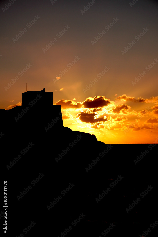 Obraz premium Striking silhouette of ancient fortress ruins on hilltop against fiery orange sunset sky, dramatic Malta landscape at dusk