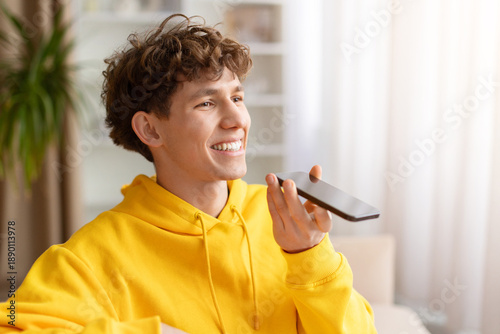 Tableau sur toile Young man in yellow hoodie using smartphone speakerphone at home, smiling during